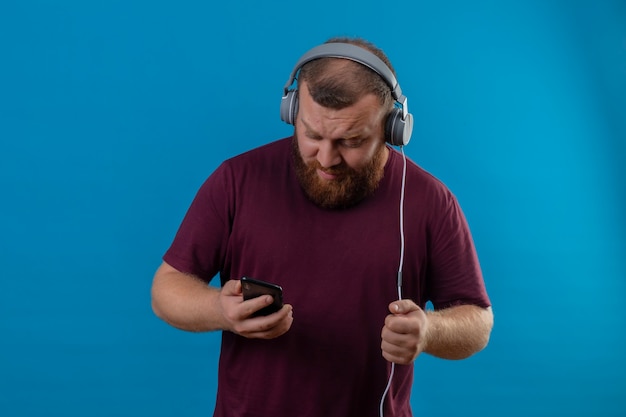 Young bearded man in brown t-shirt with headphones holding mobile phone searching favorite song  