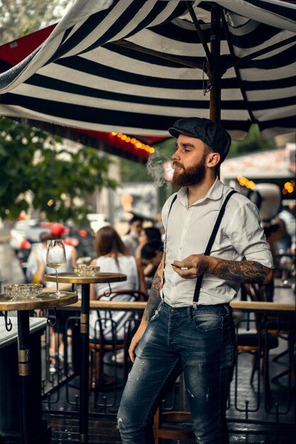 Young bearded man in a cafe on the street smokes a cigarette. Romantic guy in a white shirt cap and suspenders in the city. Peaky Blinders. old fashionable retro.