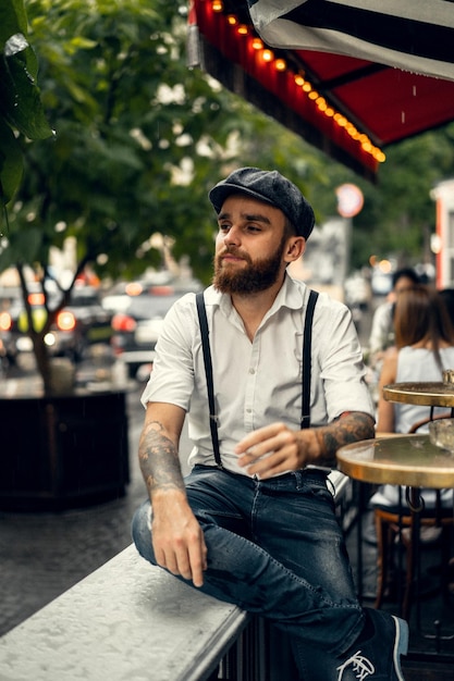 Young bearded man in a cafe on the street smokes a cigarette. Romantic guy in a white shirt cap and suspenders in the city. Peaky Blinders. old fashionable retro.