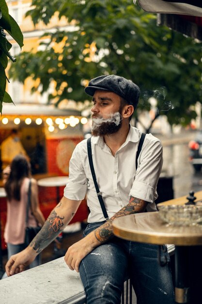 Young bearded man in a cafe on the street smokes a cigarette. Romantic guy in a white shirt cap and suspenders in the city. Peaky Blinders. old fashionable retro.