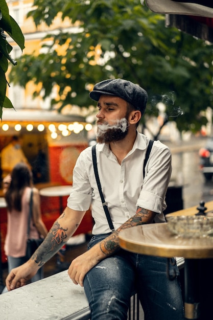 거리에 있는 카페에서 수염 난 젊은이가 담배를 피우고 있습니다. 도시에서 흰색 셔츠 모자와 멜빵에 로맨틱 남자. 피키 블라인더스. 오래 된 유행 복고풍입니다.