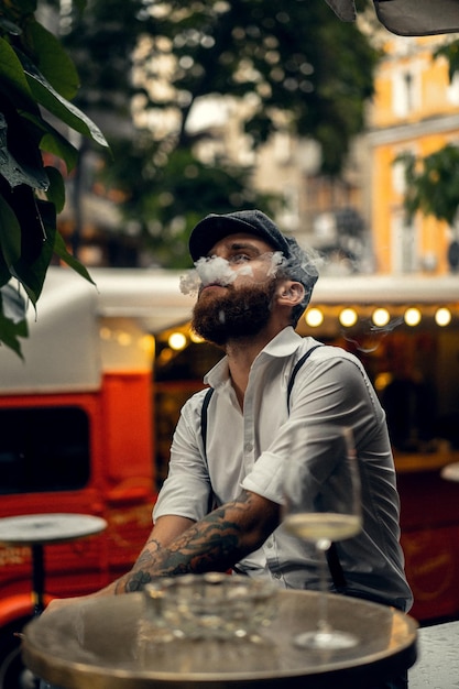 通りのカフェで若いひげを生やした男はタバコを吸います。街の白いシャツのキャップとサスペンダーのロマンチックな男。ピーキーブラインダーズ。古いファッショナブルなレトロ。