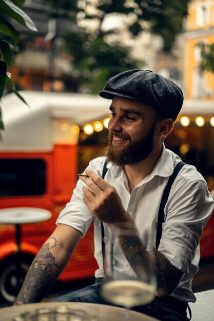 Young bearded man in a cafe on the street smokes a cigarette. Romantic guy in a white shirt cap and suspenders in the city. Peaky Blinders. old fashionable retro.
