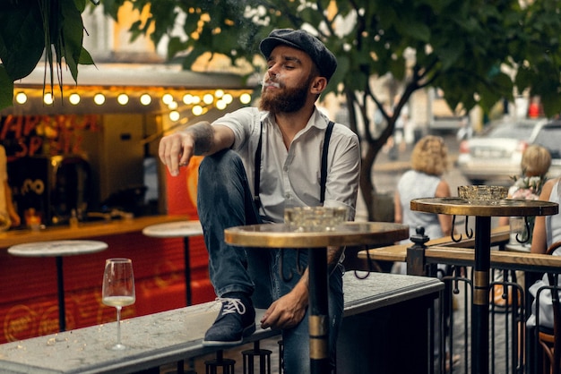 Young bearded man in a cafe on the street with a glass of wine. Romantic guy in a white shirt cap and suspenders in the city. Peaky Blinders. old fassion retro.