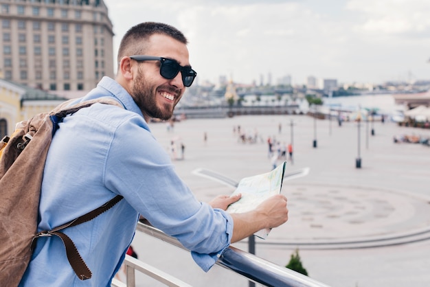 Young bearded man carrying backpack and holding map standing near railing