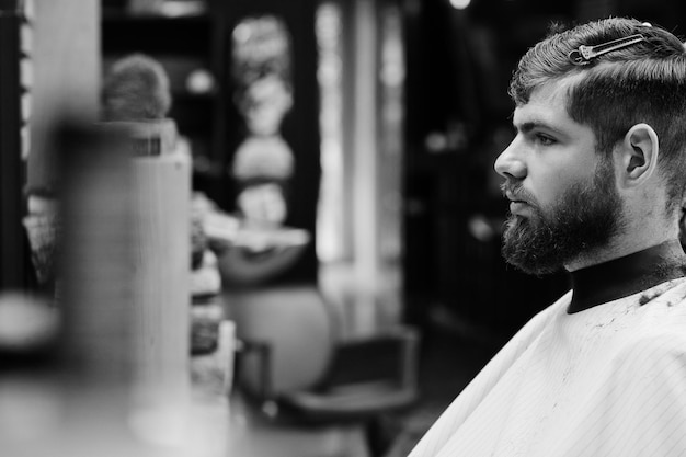 Free photo young bearded man getting haircut by hairdresser while sitting in chair at barbershop barber soul