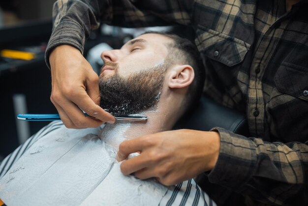 理髪店で美容師によってまっすぐなエッジのかみそりで剃られている若いひげを生やした男
