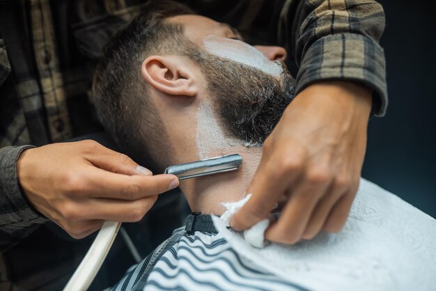 Young bearded man getting shaved with straight edge razor by hairdresser at barbershop