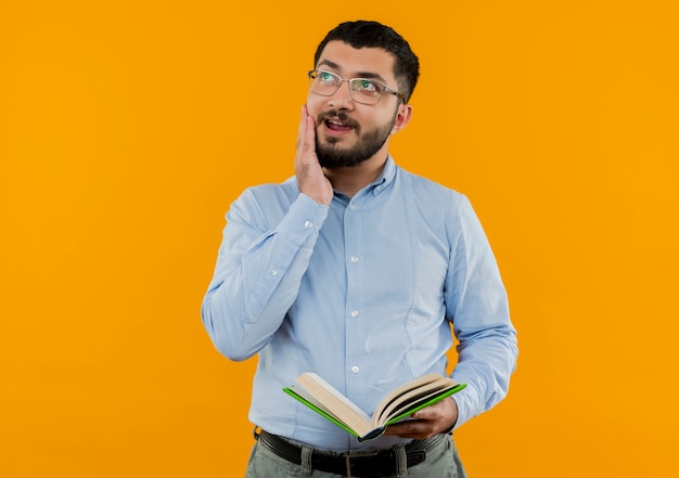 Young bearded man in glasses and blue shirt holding book looking up puzzled 