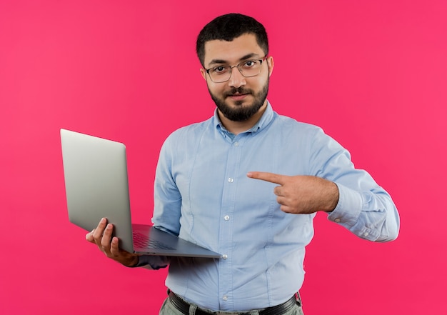 Young bearded man in glasses and blue shirt holding laptop pointign with index finger at it smiling confident 