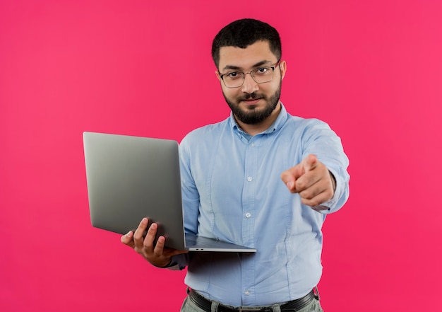 Молодой бородатый мужчина в очках и синей рубашке держит ноутбук, указывая указательным пальцем на камеру с уверенным серьезным выражением лица