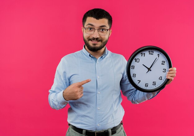 Young bearded man in glasses and blue shirt holding wall clock pointing with index finger at it smiling with happy face 