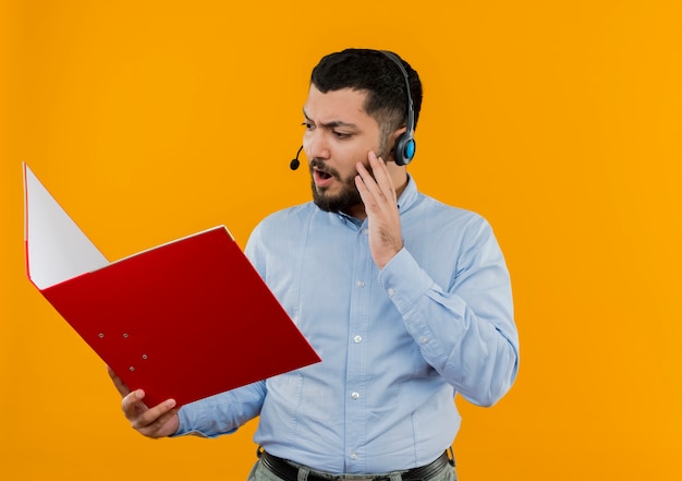 Young bearded man in glasses and blue shirt with headphones with microphone holding big folder looking at it surprised and amazed 