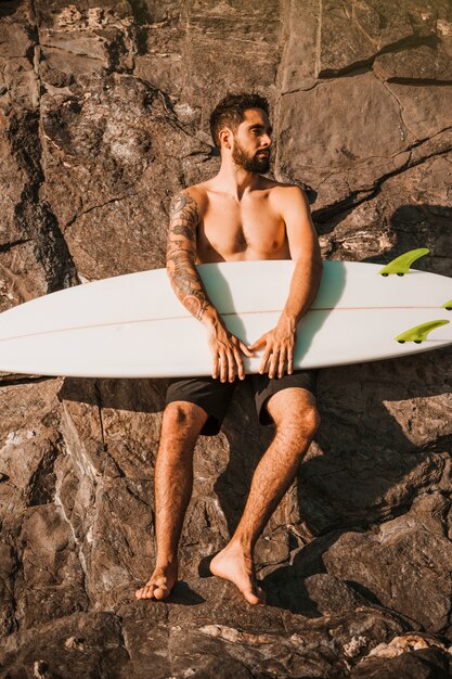 Young bearded man holding surf board near stones