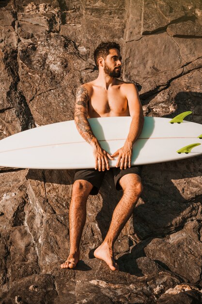 Молодой бородатый мужчина держит доску для серфинга возле камней