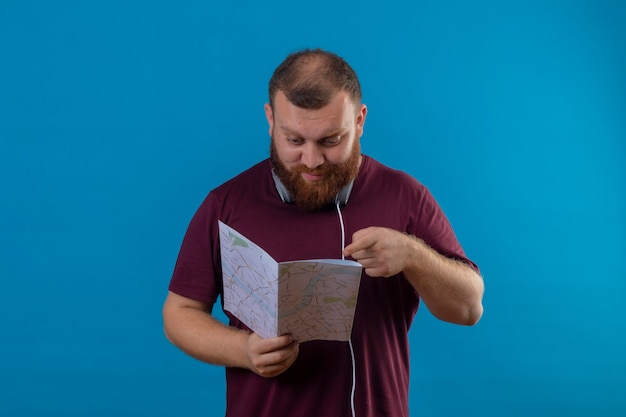 Молодой бородатый мужчина в коричневой футболке с наушниками на шее держит карту, глядя на нее, указывая пальцем с серьезным лицом
