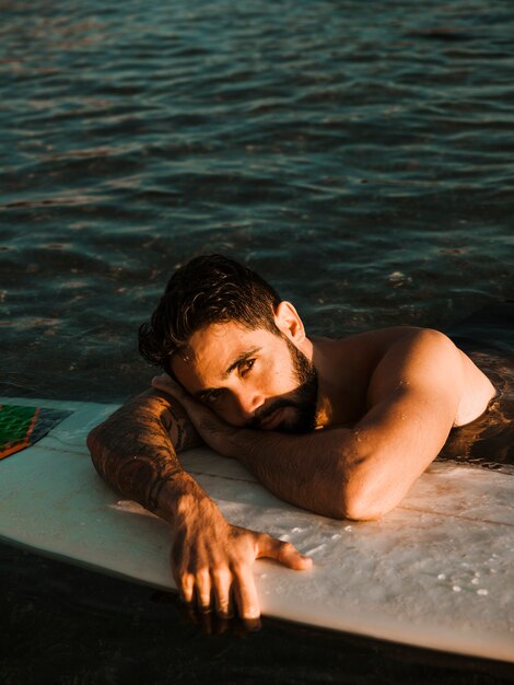 Young bearded man lying on surf board in sea near coast