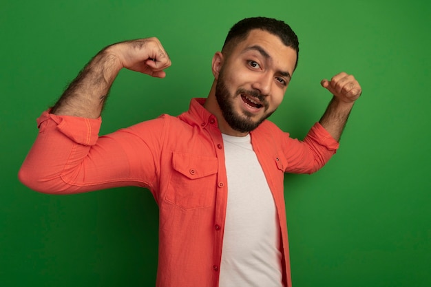 Young bearded man in orange shirt clenching fists happy and excited rejoicing his success standing over green wall