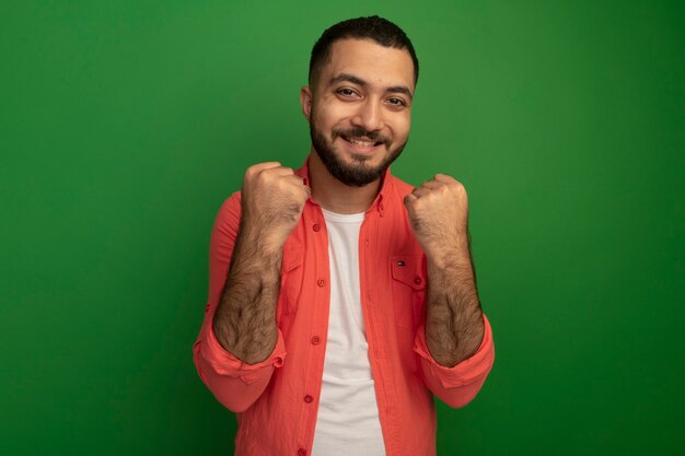 Young bearded man in orange shirt clenching fists happy and excited rejoicing his success standing over green wall