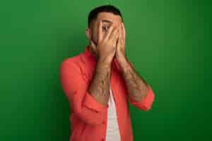 Free photo young bearded man in orange shirt covering face with palms looking  through fingers being scared standing over green wall