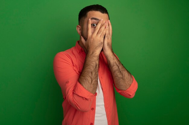 Молодой бородатый мужчина в оранжевой рубашке, закрывая лицо ладонями, глядя сквозь пальцы, испугался, стоя над зеленой стеной