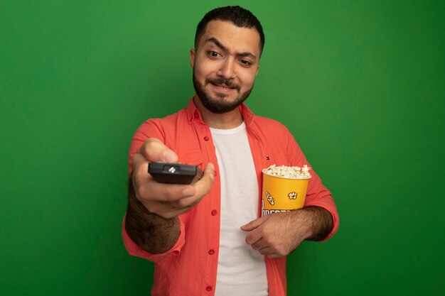 Young bearded man in orange shirt holding bucket with popcorn using tv remote looking at it confused standing over green wall