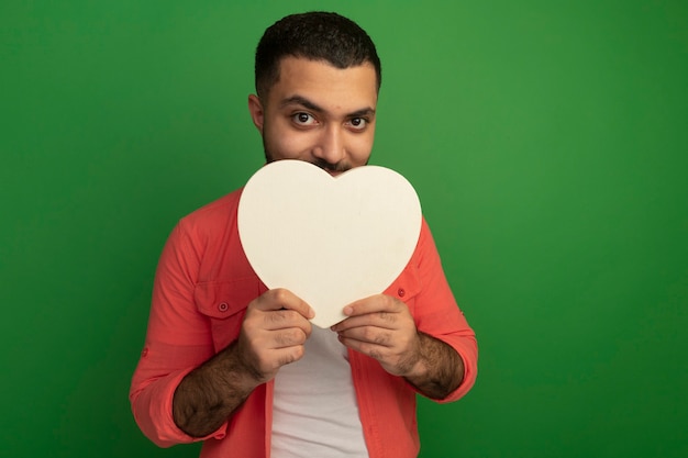 Young bearded man in orange shirt holding cardboard heart looking  smiling slyly standing over green wall