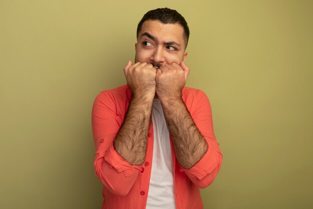 Young bearded man in orange shirt looking aside stressed and nervous biting nails standing over light wall