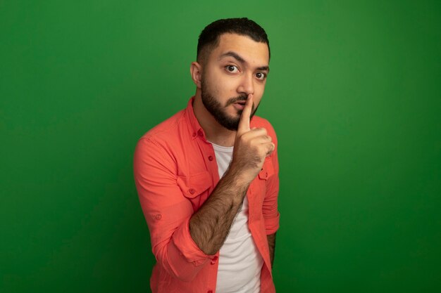 Молодой бородатый мужчина в оранжевой рубашке делает жест молчания с пальцем на губах, стоя над зеленой стеной