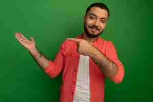 Free photo young bearded man in orange shirt presenting something with arm pointing with index finger to the side smiling confident standing over green wall