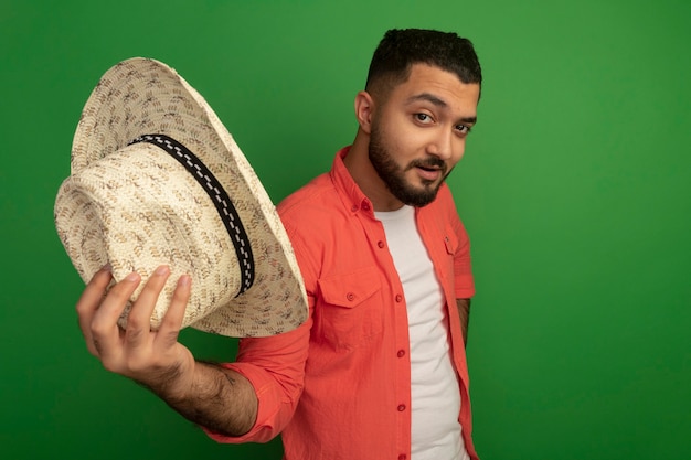 Free Photo young bearded man in orange shirt showing his summer hat  smiling confident standing over green wall