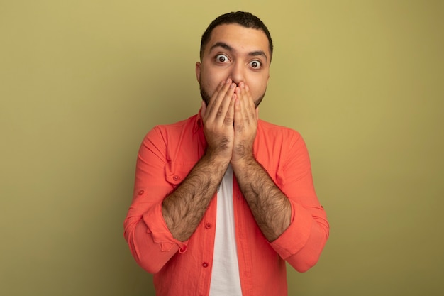 Young bearded man in orange shirt  surprised covering mouth with hands standing over light wall