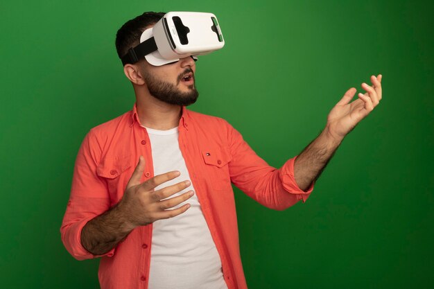 Молодой бородатый мужчина в оранжевой рубашке с очками виртуальной реальности жестикулирует руками, стоящими над зеленой стеной