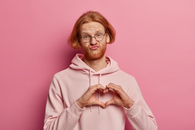 Young bearded man pouts lips and makes heart gesture on his chest, wears casual sweatshirt, expresses affection, sympathy and love, has ginger hair, being in love with woman, isolated on pink