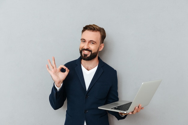 Young bearded man in suit looking camera while holding laptop computer isolated