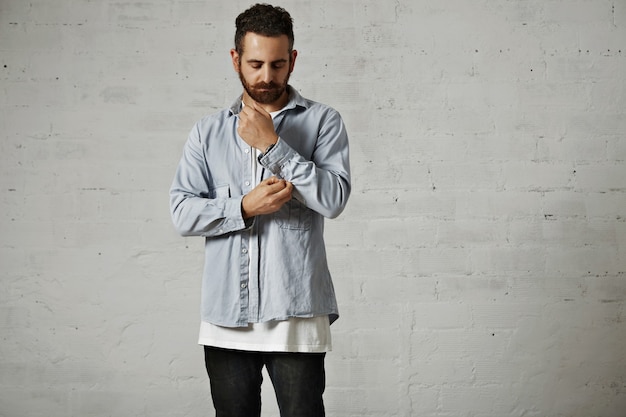 Free photo young bearded man wearing a light blue denim shirt and black jeans unbuttoning his shirt sleeve on white brick wall