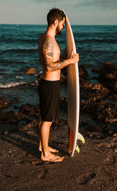Young bearded man with surf board on shore near water