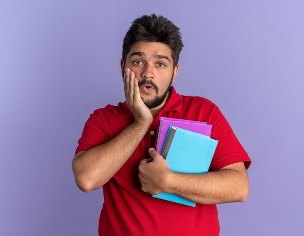 Young bearded student guy in red polo shirt holding books  amazed and surprised standing over blue wall