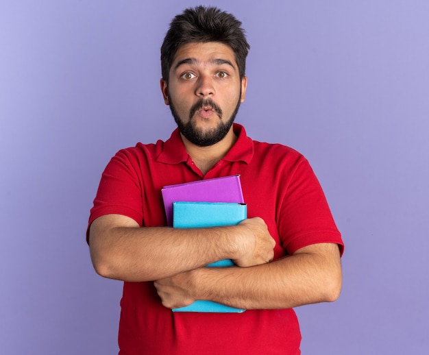 Young bearded student guy in red polo shirt holding books looking amazed and surprised standing