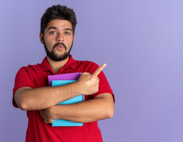 Young bearded student guy in red polo shirt holding books looking surprised pointing with index finger to the side standing