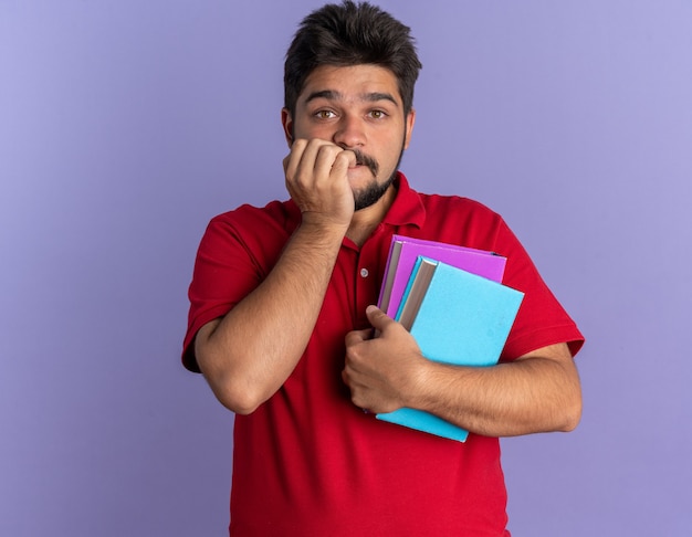 Молодой бородатый студент-парень в красной рубашке поло держит книги в стрессе и нервно кусает ногти, стоя у синей стены