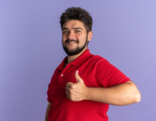 Young bearded student guy in red polo shirt looking happy and positive smiling showing thumbs up standing