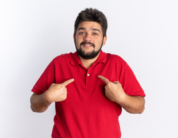 Молодой бородатый студент-парень в красной рубашке поло выглядит счастливым и удивленным, указывая указательными пальцами на себя стоящего