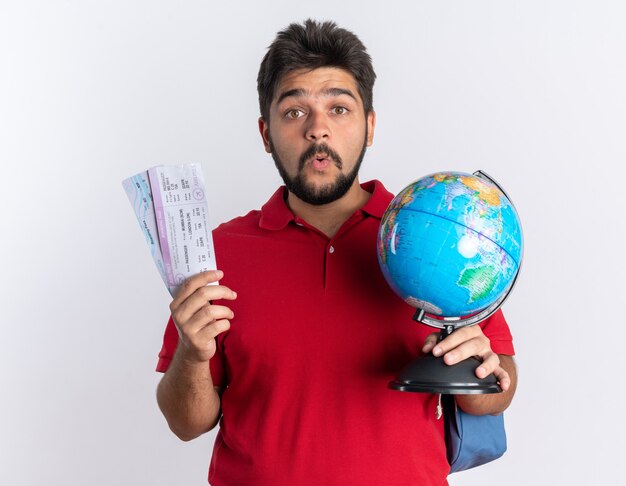 Молодой бородатый студент-парень в красной рубашке поло с рюкзаком с билетами на самолет и земным шаром, выглядящим удивленным стоя