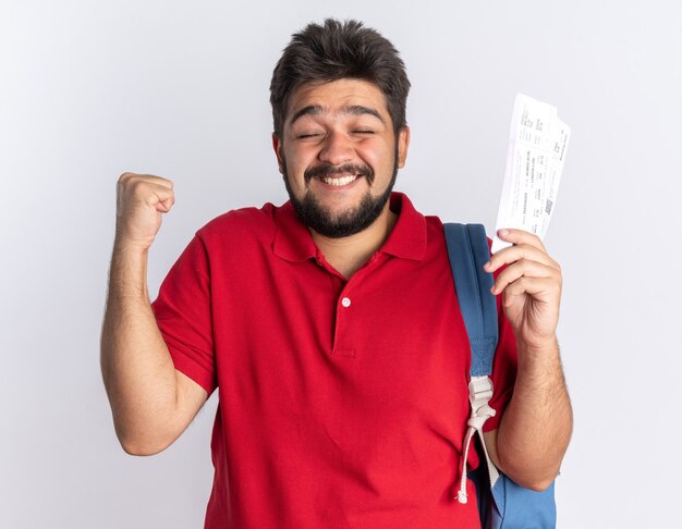 Young bearded student guy in red polo shirt with backpack holding airline tickets happy and excited clenching fist standing