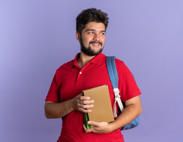 Young bearded student guy in red polo shirt with backpack holding books looking aside with smile on happy face standing