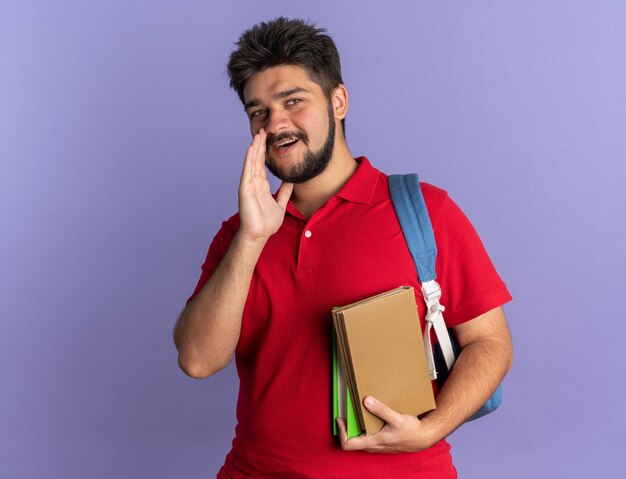 Ragazzo giovane studente barbuto in maglietta polo rossa con zaino in possesso di libri che guarda l'obbiettivo sussurrando un segreto con la mano vicino alla bocca in piedi felice e positivo su sfondo blu