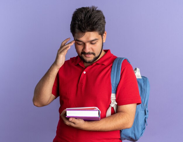 赤いポロシャツを着た若いひげを生やした学生の男が本を持ってバックパックを見て、自信を持って幸せで、青い壁の上に前向きに立っている