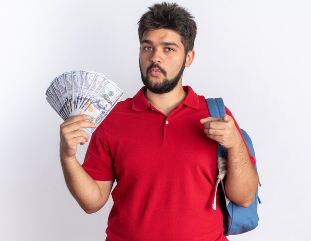 Free photo young bearded student guy in red polo shirt with backpack holding cash looking confident pointing with index finger standing