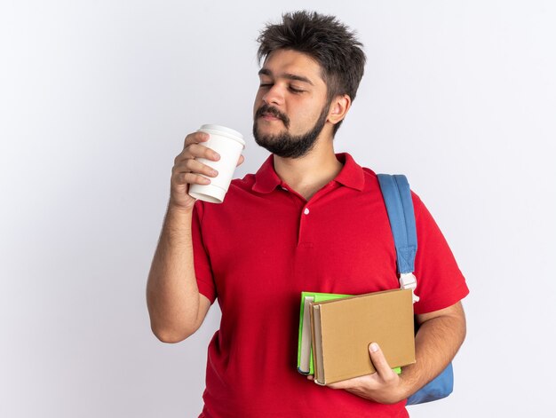 Young bearded student guy in red polo shirt with backpack holding notebooks and coffee cup looking confident smiling standing over white background
